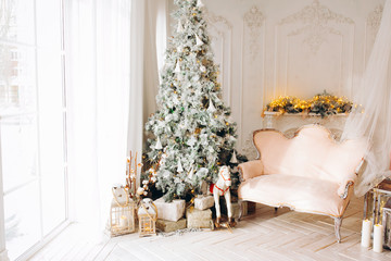 classical interior of a white room with a decorated fireplace, luxury Christmas tree, garlands, candles, lanterns, gifts. day. pink sofa pastel colors