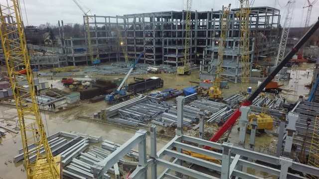 An aerial view of a busy construction site on a dirty puddly ground. It is filled with construction cranes, multi storey building frames and metal beams. There is a misty urbanscape among leafless