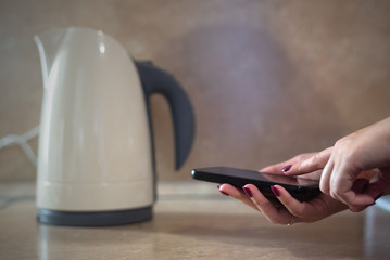 Fototapeta premium Woman is warming up a water in the kettle by a mobile phone with smart home application concept.