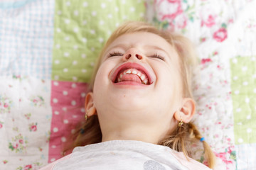 Pretty happy little smiling girl with blond hair lying in bed on a quilted blanket.