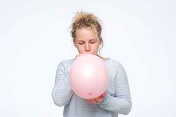 Young caucasian curly woman in gblue sweateris blowing pink balloon preparing for birthday party....