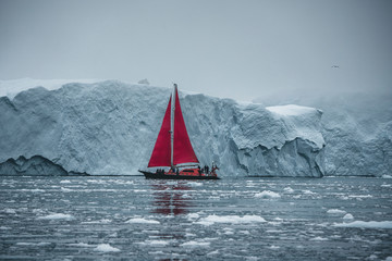 Beautiful red sailboat in the arctic next to a massive iceberg showing the scale. Cruising among...