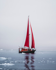 Naklejka premium Beautiful red sailboat in the arctic next to a massive iceberg showing the scale. Cruising among floating icebergs in Disko Bay glacier during midnight sun season of polar summer Ilulissat, Disko Bay