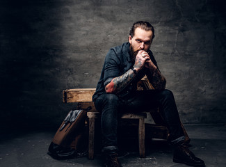 Obraz na płótnie Canvas Pensive bearded man with tattooed arms is sitting at dark photo studio.