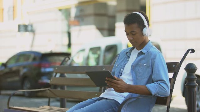 Mixed-race guy listening to music in headphones and scrolling tablet, resting