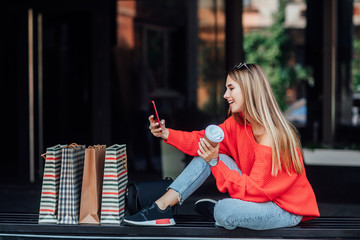 Beautifulblonde woman sitting on a street and surrounded by shopping bags and looking on phone.