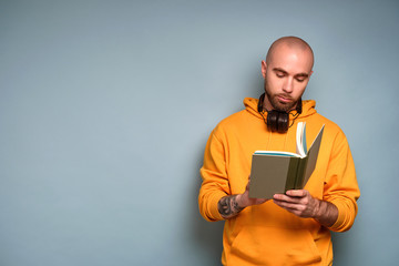 A bearded guy in a yellow sweatshirt stands on a blue background with headphones on his neck, looks into an open book in his hands