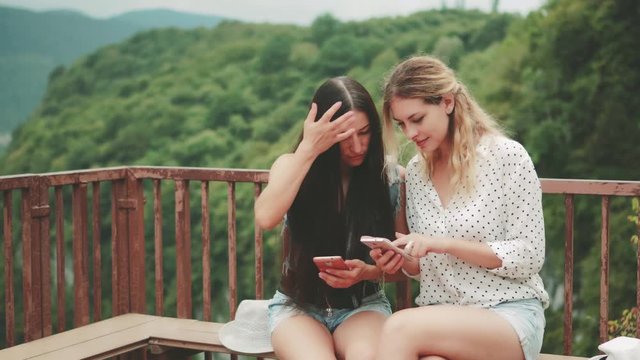 Two attractive women are sitting on a bench on top with a picturesque view of the mountains and chatting. Girl shows a photo to her friend on a mobile phone. Dependence on modern gadgets.