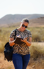 young woman with sunglasses and backpack writing notes in her notebook in nature