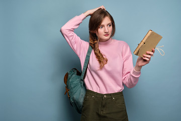 A girl in a pink sweater with a backpack stands on a blue background and holds a book, looking puzzled at her, scratching her head