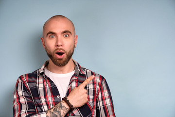 A bearded guy in a plaid shirt stands on a blue background, opening his mouth and eyes in surprise, shows a finger to the side