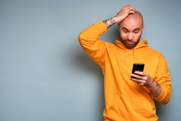 A bearded guy in a yellow sweatshirt stands on a blue background and looks in horror at the smartphone with his hand on his head