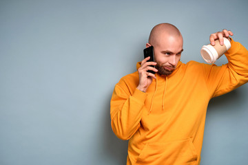 A guy with a beard in a yellow sweatshirt is standing with a phone near his ear looking displeased at the cup tilting it