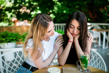 Young girl talking with her upset friend and holding her shoulders in cafe.