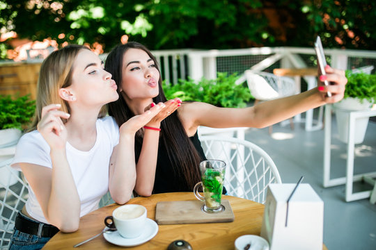 Cheerful female friends blow kiss taking selfie picture in cafe. Two beautiful young women smiling and looking to the camera in smartphone. Friendship concept