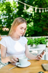 Beautiful young woman sits at a table in a cafe with a cup of coffee and uses the phone