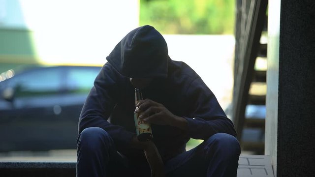 Troubled teenager drinking beer sitting on steps in gateway, lost in life