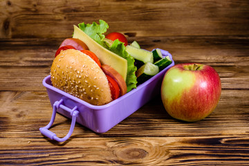 Ripe apple and lunch box with hamburger, cucumbers and tomatoes on wooden table