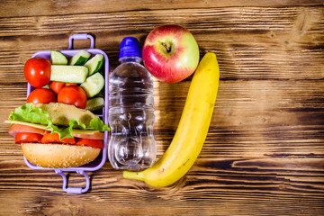 Ripe apple, banana, bottle of water and lunch box with hamburger, cucumbers and tomatoes on wooden table. Top view