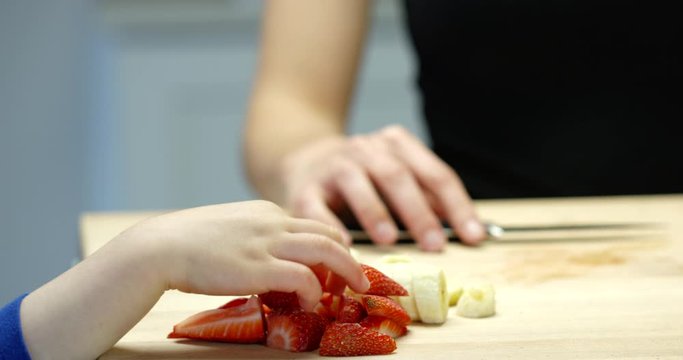 Child hands pick strawberries off of cutting board in kitchen while mother cuts up fruite - close up on hands