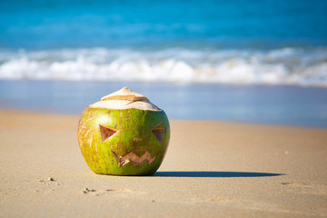 Green coconut with a face carved on it, like a halloween pumpkin. Lies in the sand on a tropical beach on a background of waves. Holiday trip concept.