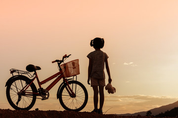 Silhouette of little girl standing with bike at sunset background