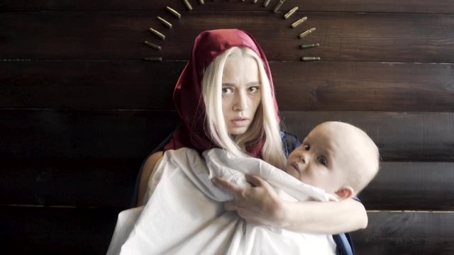 Woman with the covered head, mother with a baby in her arms standing against wooden wall with an arc of bullets, religion and war concept. Footage. The image of the virgin Mary and a Jesus.