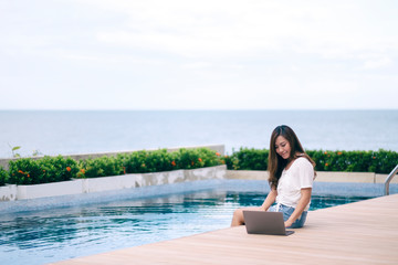 A beautiful asian woman using and typing on laptop computer while sitting by swimming pool