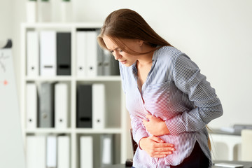 Young woman suffering from abdominal pain in office - obrazy, fototapety, plakaty