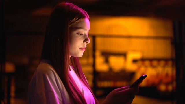 Young woman scrolling web-pages on smartphone waiting for friends near club
