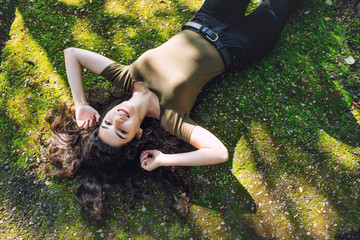 Young adult beautiful brunette girl in casual clothes happy lying on green moss in Sunny Park
