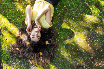 Young adult beautiful brunette girl in casual clothes happy lying on green moss in Sunny Park