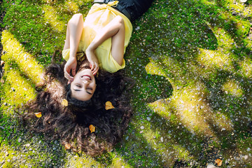 Young adult beautiful brunette girl in casual clothes happy lying on green moss in Sunny Park