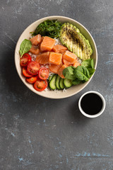 Organic poke bowl with salmon, avocado and vegetables on dark background