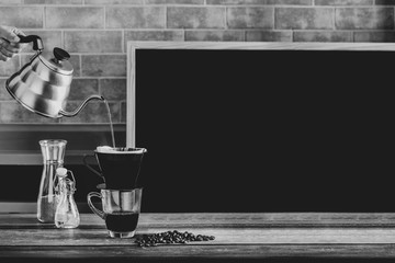 Hand holding a teapot for drip brewing coffee by pouring down hot water on coffee beans and filter on a cup of coffee at a coffee shop, brick wall and a blackboard in background, black and white tone.