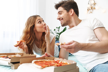 Image of lovely happy couple lying on bed at home and eating pizza