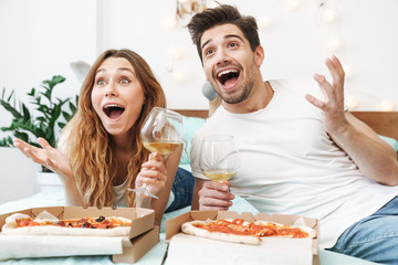 Image of joyful couple drinking wine and eating pizza on bed at home