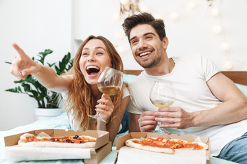 Image of cheerful couple drinking wine and eating pizza on bed at home