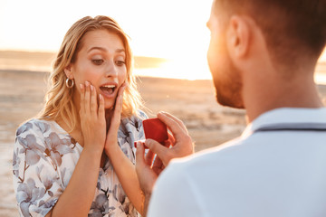 Photo of unshaven man making proposal to his amazed woman with ring while walking on sunny beach - obrazy, fototapety, plakaty