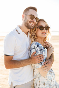 Photo of smiling caucasian couple man and woman looking aside and hugging while walking on sunny beach