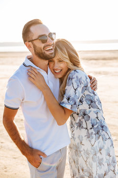 Photo of pleased pretty couple laughing and hugging while walking on sunny beach