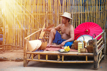 An Asian man do not wear shirts sitting on a bamboo carriage, Bamboo bed for sitting with bamboo hat, red umbrella and Yellow metal Tiffin,thai food carrier.