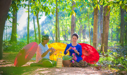 Two Asian women sitting in forest and Arrange pink lotus flowers, water lilly to pay respect to the Buddha with yellow, red umbrellas and tree background.