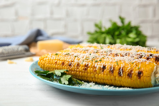 Plate of grilled corn cobs with grated cheese on white wooden table, closeup