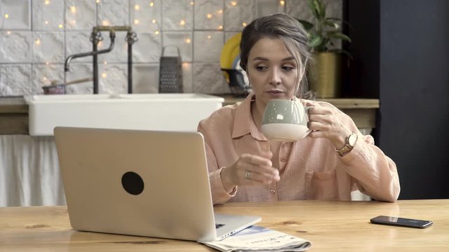 Woman working from home using laptop In the kitchen. Stock footage. Cute brunette woman drinking tea from grey and beige mug, working on laptop, freelance concept.