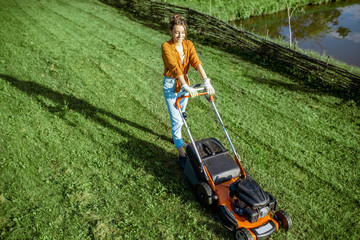 Beautiful young woman cutting grass with gasoline lawn mower, gardening on the backyard in the countryside, view from above