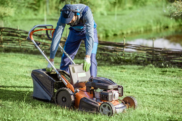 Fototapeta premium Professional gardener in protective workwear getting grass basket from the lawn mower while cutting grass on the backyard