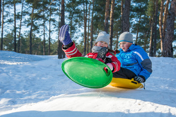 In winter, in the forest on a bright sunny day, brother and sister ride plastic plates from the mountain.