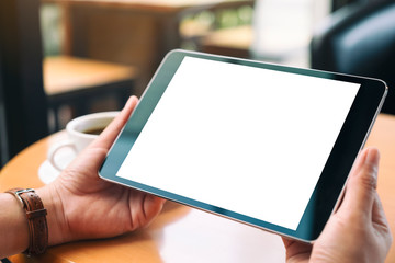 Mockup image of hands holding black tablet pc with blank white screen with coffee cup on wooden table