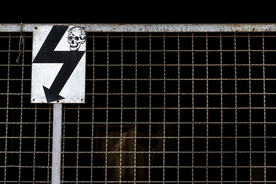 Restricted area and safety zone photo. Old electric sign and skull picture shown on white metal plate on dirty steel fence and wire mesh in front of high voltage electricity machine background.